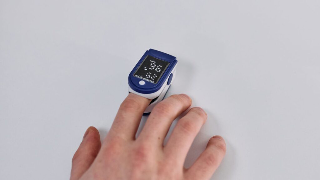 A hand resting on a light gray surface with a blue fingertip pulse oximeter clipped to one finger, its screen showing an oxygen reading