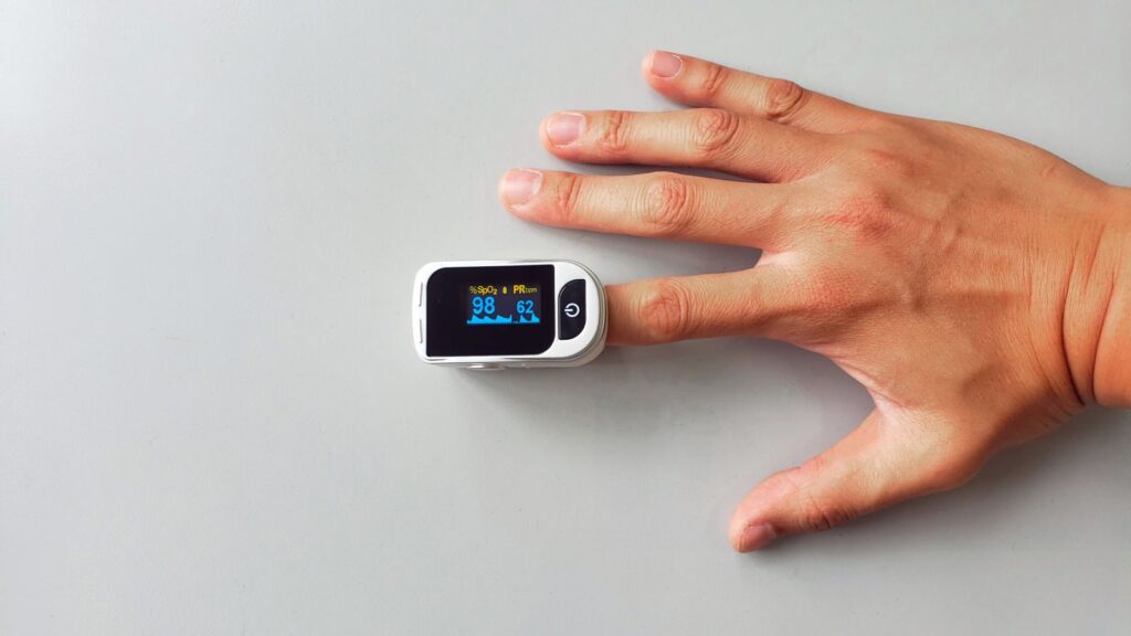 Close-up of a hand wearing a finger pulse oximeter that shows a health reading